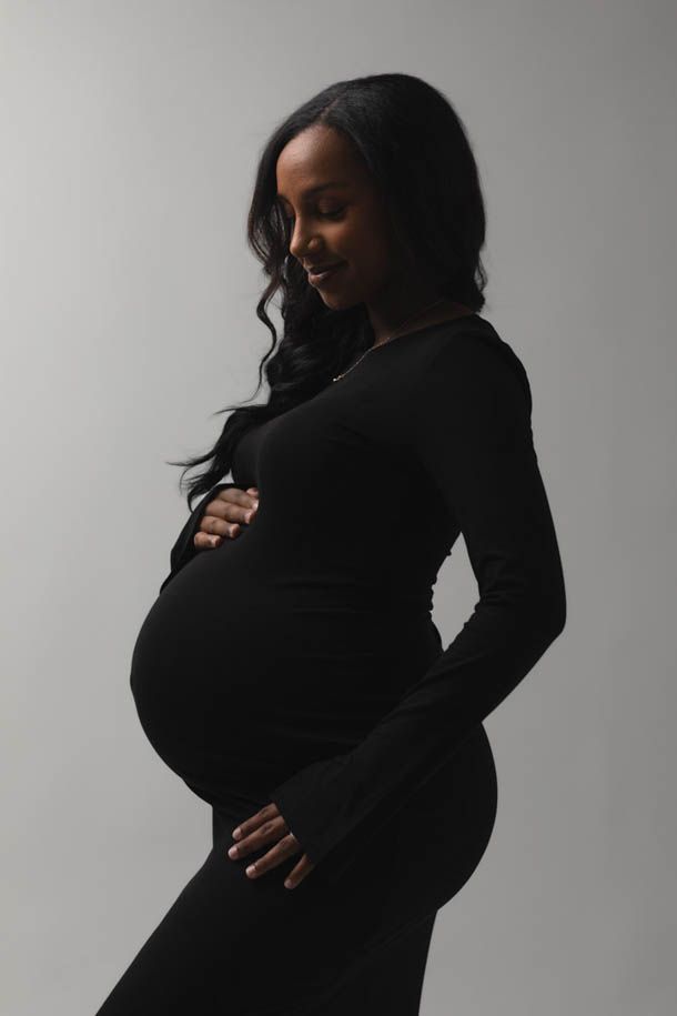 Mor i sort kjole med gravidmage poserer foran hvit bakgrunn i fotostudio