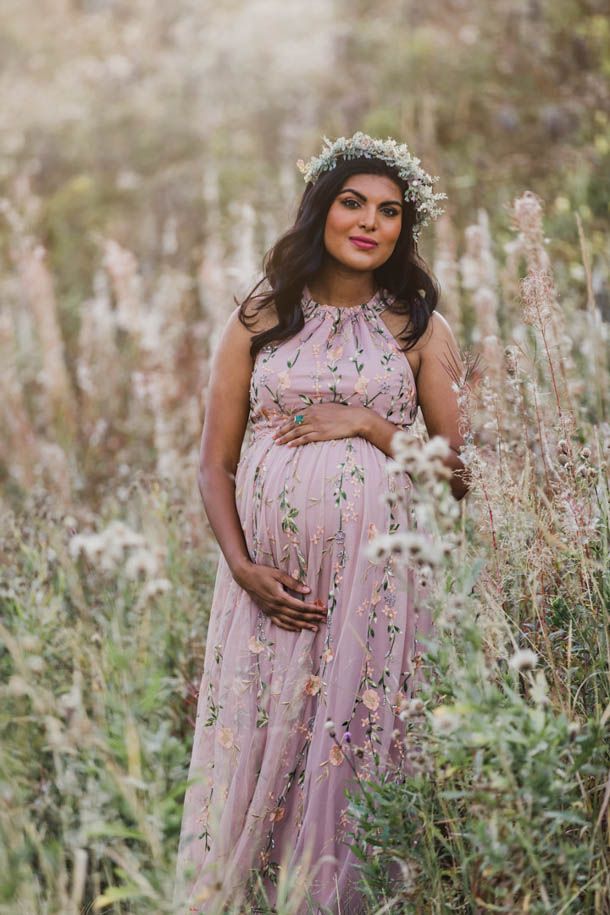 Gravidfotografering - Dame med blomsterkrans i h&aring;ret og blomstrete rosa kjole holder p&aring; magen utend&oslash;rs i h&oslash;yt gress