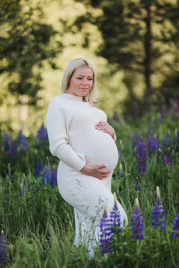 Gravidfotografering - Gravid dame med lyst h&aring;r i hvit kjole holder p&aring; magen i en eng med bl&aring; lupiner