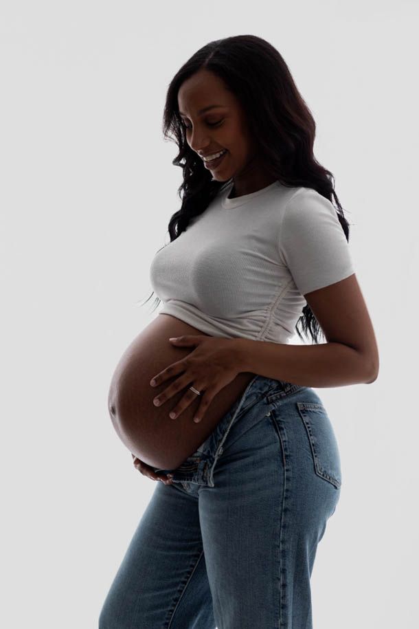 Dame med gravidmage, jeans og hvit topp poserer avslappet foran hvit bakgrunn i studio. 