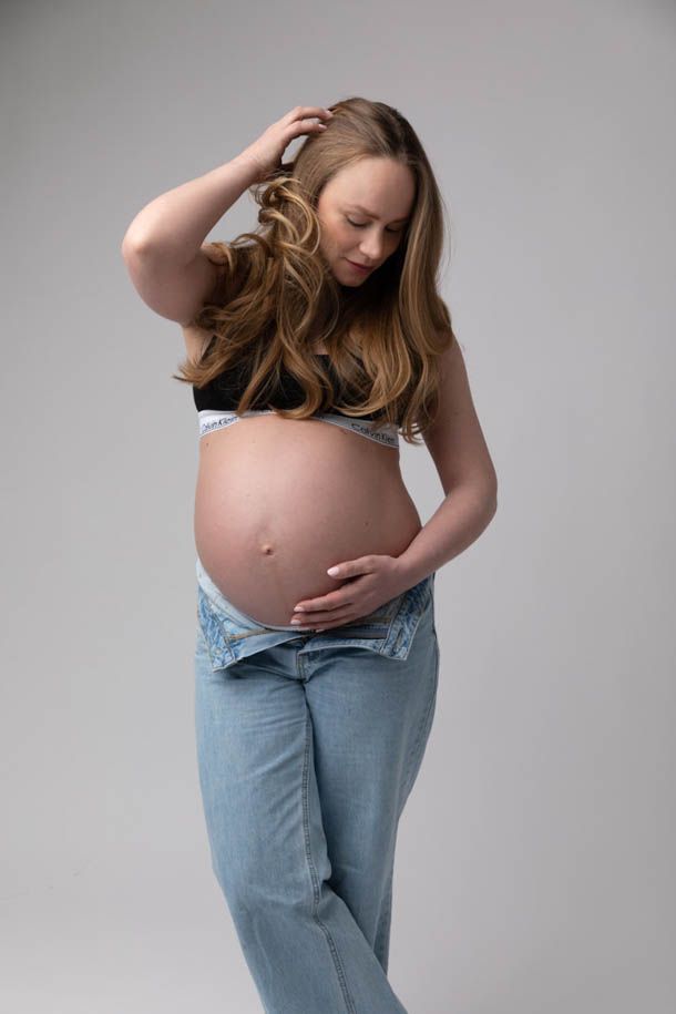 Kvinne med lyst h&aring;r og bl&aring; jeans holder p&aring; gravidmagen og fjerner h&aring;ret fra ansiktet med andre h&aring;nden. 