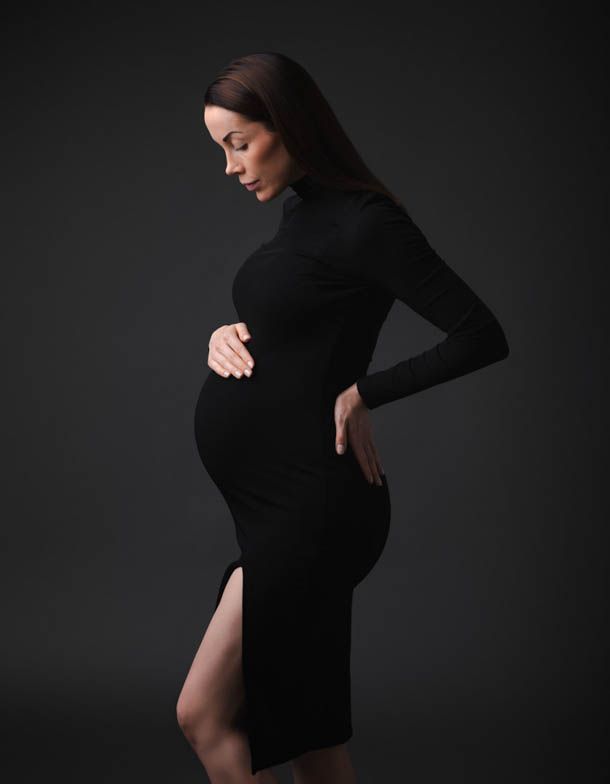 Gravidfotografering - gravid dame i sort kjole forran sort bakgrunn holder en h&aring;nd p&aring; magen og ser ned