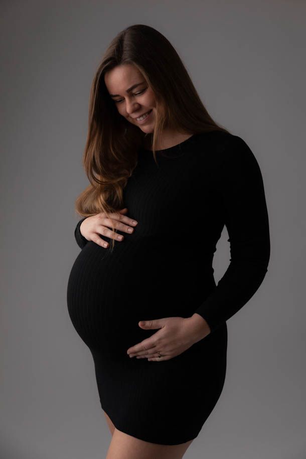 Gravidfotografering - Gravid dame som smiler i sort kjole, holder rundt magen og titter ned p&aring; magen i studio. 