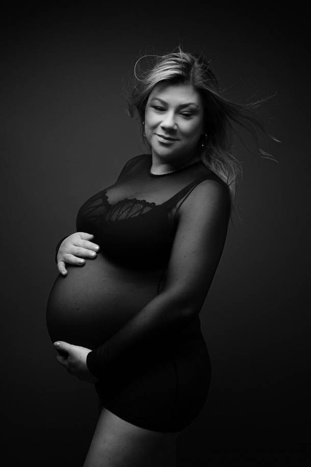 Gravidfotografering - dame i sort kjole mot sort bakgrunn poserer med vind i h&aring;ret