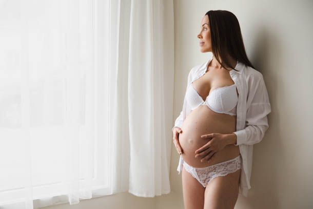 Gravid dame med hvit skjorte holder p&aring; magen og titter ut av vindu