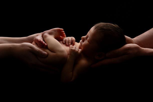 Nyf&oslash;dtfotografering - Mamma og pappas hender holder nyf&oslash;dt baby ifra hver sin side med sort bakgrunn og motlys