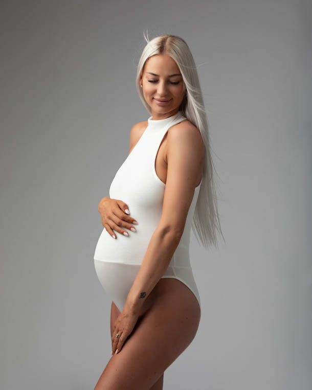Gravidfotografering - Dame i hvitt body med langt h&aring;r foran hvit bakgrunn titter ned og smiler
