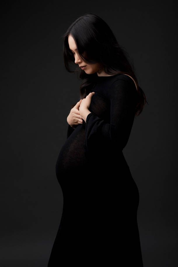 Gravidfotografering - Dame med m&oslash;rkt h&aring;r i sort kjole forran sort bakgrunn holder begge hendene til brystet og titter ned p&aring; magen